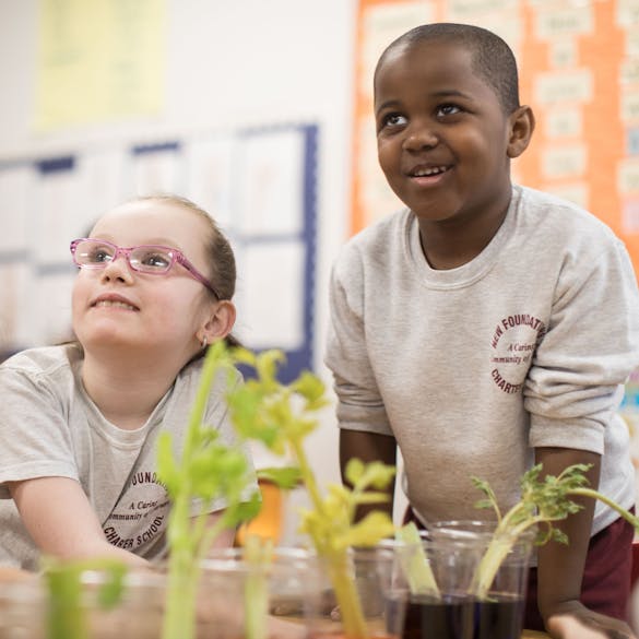  A young male and female student look up from behind plants growing in cups inside the classroom 