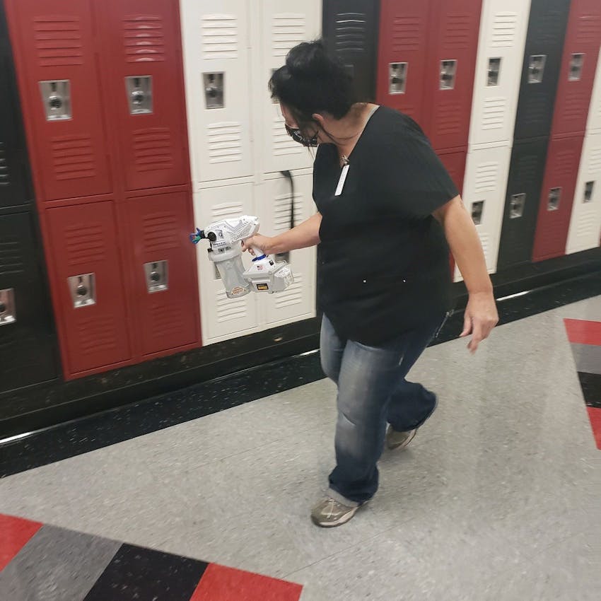 Female custodian wearing mask and spraying sanitizer on lockers in hallway.