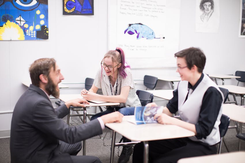 Male teacher working with two smiling students at desks with novels open.
